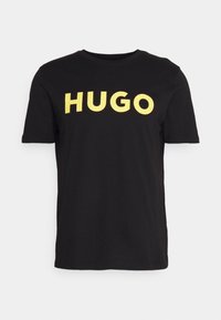 Camiseta de algodón negra con un llamativo texto amarillo "HUGO" en el pecho, cuello redondo y mangas cortas. Diseño simple y moderno.