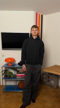 Jonge man in een zwarte hoodie en grijze broek staat in een kamer met aan de muur gemonteerde tv, een oranje lamp op een plank, een basketbal en een koffer met stickers.