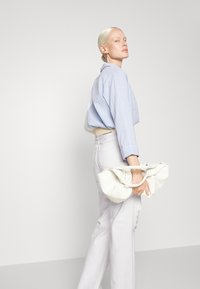 Blusa oversized às riscas azuis e brancas, faixa na cintura bege, calças largas de cor clara, segurando uma mala de couro cream plissada.