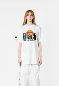 Camiseta blanca de corte oversize hecha de algodón, con un gráfico texturizado de un animal marrón y texto negro que dice "Black". Tiene mangas cortas y un cuello redondo.