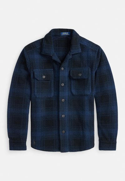 Plava i crna flanelasta košulja na kopčanje s ovratnikom i dva džepa na prsima s kopčama, položena ravno na bijelu podlogu.