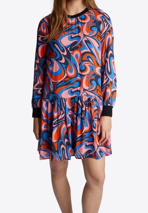 Kobieta ubrana w sukienkę do kolan z długimi rękawami, marszczoną w talii, z wirującym abstrakcyjnym wzorem w kolorach niebieskim, różowym, czerwonym i czarnym.