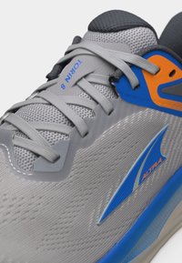 Γκρι και μπλε αθλητικό παπούτσι με διχτυωτό επάνω μέρος, γκρι κορδόνια και πορτοκαλί-μπλε στήριγμα φτέρνας με ετικέτα "TORIN 8" και λογότυπο "ALTRA" στην πλευρά.