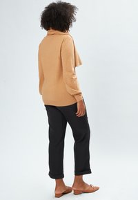 Beige Pullover mit Kapuze und gepunkteter Textur, kombiniert mit schwarzen Hosen mit umgeschlagenem Saum und braunen Slippern.
