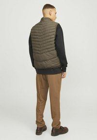 Olivgrüne Steppweste mit Zickzack-Quiltmuster, getragen über einem schwarzen Sweatshirt, kombiniert mit braunen Hosen und dunklen Schuhen.