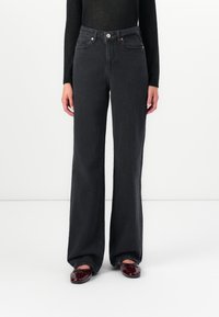 Άτομο που φοράει μαύρα ψηλόμεσα φαρδιά τζιν, μαύρη μπλούζα με μακριά μανίκια και σκούρα κόκκινα γυαλιστερά μοκασίνια, στεκόμενο μπροστά σε λευκό φόντο.