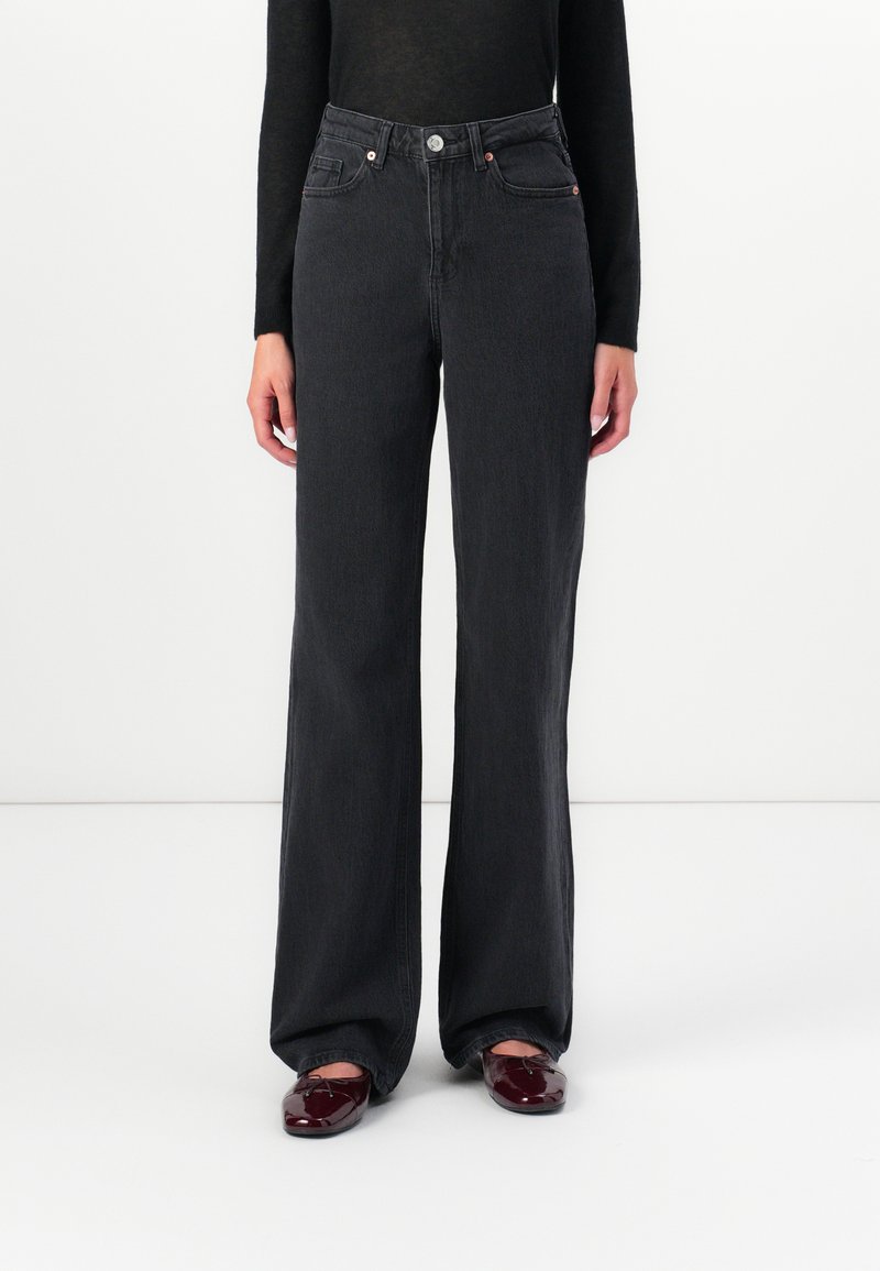 Άτομο που φοράει μαύρα ψηλόμεσα φαρδιά τζιν, μαύρη μπλούζα με μακριά μανίκια και σκούρα κόκκινα γυαλιστερά μοκασίνια, στεκόμενο μπροστά σε λευκό φόντο.