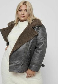 Chaqueta de piel sintética gris con un cuello de borreguito marrón, bolsillos con cremallera y un diseño recortado, llevada sobre un vestido de suéter de punto blanco.