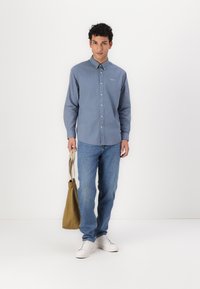 Cămașă albastră, cu mâneci lungi și nasturi; stofă moale și guler. Blugi de culoare albastru deschis. Adidași albi. Ținând o geantă de tote din material bej.