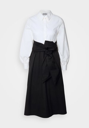 Camicia bianca con colletto e maniche a sbuffo lunghe, abbinata a una gonna nera e una cintura di tessuto annodata in vita, realizzata in cotone morbido.
