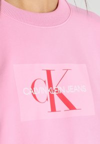 Felpa rosa con scollo a costine, con un grande logo rettangolare in rosso e bianco che dice "CALVIN KLEIN JEANS" e "CK."