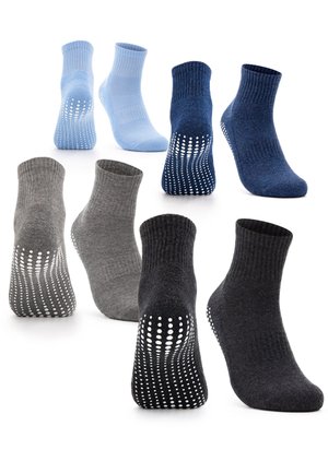 Beine tragen schwarze rutschfeste Socken mit weißen Griffen, ein Fuß flach auf dem Boden, der andere auf den Zehen erhoben vor einfachem Hintergrund.