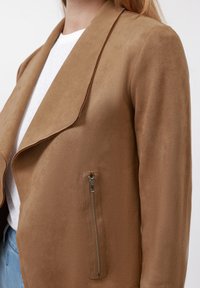 Gros plan sur une personne portant une veste en daim marron avec une poche zippée en métal, une chemise blanche et un jean bleu sur fond blanc.