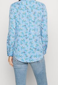 Blusa azzurro chiaro con motivo floreale, maniche lunghe, orlo arrotondato e dettagli con bottoni sul retro. Il tessuto appare morbido e texture.
