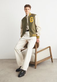 Bombe en vert olive et crème avec des rayures jaunes et un grand logo "G", associée à un pantalon crème et des mocassins noirs. Chaise en bois en arrière-plan.
