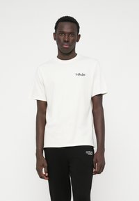 Κρέμα βαμβακερό μπλουζάκι με κοντό μανίκι, στρογγυλή λαιμόκοψη και μικρή μαύρη γραφική παράσταση στο στήθος. Φοριέται με μαύρες φόρμες.