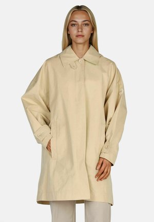 Woman stands facing forward wearing a loose beige trench coat with large collar and buttoned cuffs, hands partially in pockets.