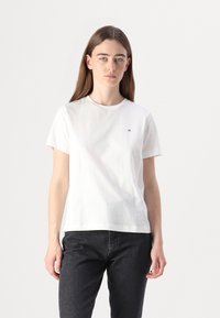 T-shirt bianco in cotone con scollo rotondo e maniche corte, con un piccolo logo a contrasto sul lato sinistro del petto. Indossato con jeans scuri.