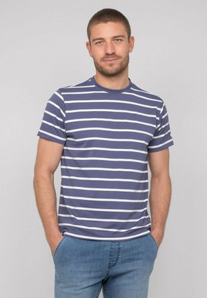 Homme aux cheveux courts debout, les mains dans les poches, portant un t-shirt rayé bleu et blanc et un jean bleu sur un fond uni.