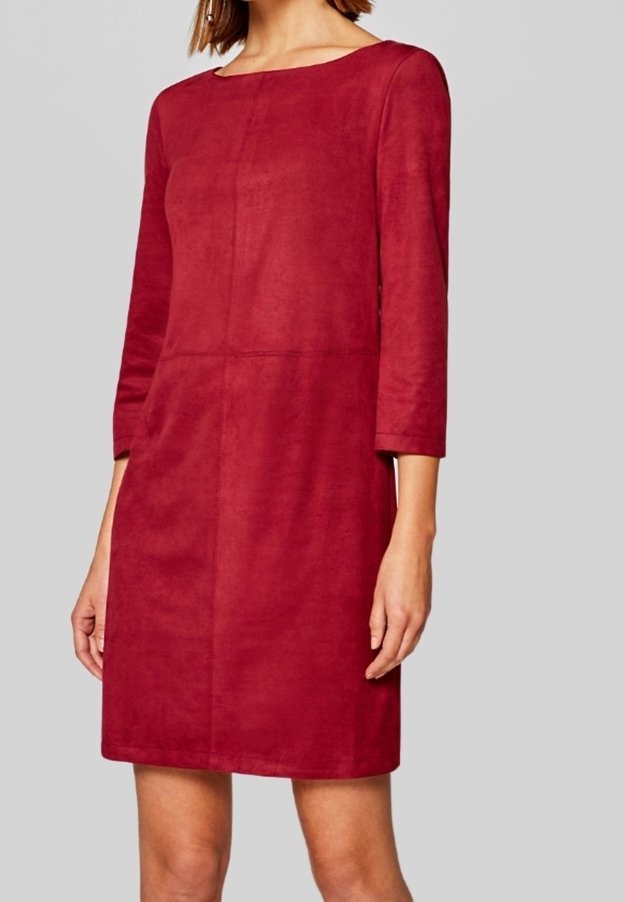 Femme portant une robe rouge longue jusqu'aux genoux, à manches longues et col bateau, sur un fond gris clair uni.