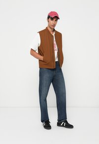 Gilet sans manches marron sur un t-shirt graphique blanc, associé à un jean bleu et des baskets noires avec des accents blancs, sur un fond uni.