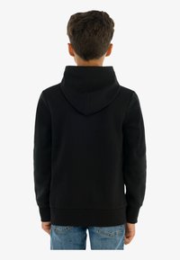 Sudadera de algodón negra con un bolsillo canguro, puños acanalados y capucha con cordón, llevada con jeans azul claro, vista desde atrás.