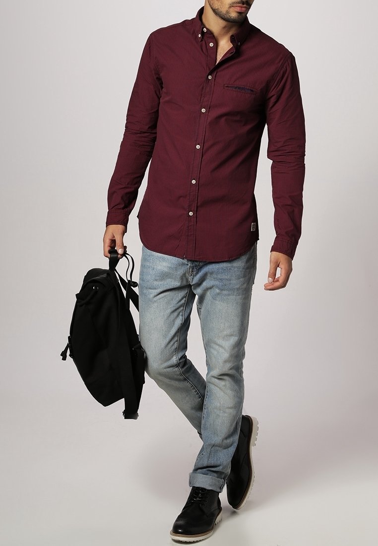 Homme marchant, portant une chemise bordeaux à boutons, un jean bleu clair, des chaussures noires et tenant un sac à dos noir par une sangle.