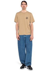 Beige cotton t-shirt with short sleeves and a small printed design on the left chest, paired with loose blue denim pants and black sneakers.