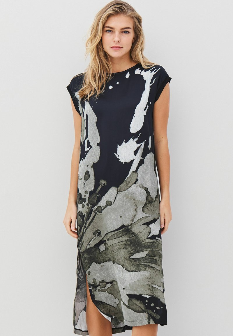 Femme aux cheveux blonds portant une robe midi noire sans manches avec un grand imprimé floral abstrait gris, posant devant un fond blanc uni.