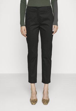 Mujer con pantalones negros ajustados hasta el tobillo, camiseta verde de canalé de manga larga y tacones marrones con punta afilada con estampado, sobre un fondo liso.