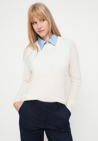 Ξανθιά γυναίκα που φοράει ένα λευκό πουλόβερ επάνω από ένα μπλε πουκάμισο με γιακά και ναυτικούς παντελόνες, με τα χέρια στις τσέπες, κοιτώντας στο πλάι.