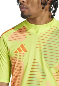 Maglietta atletica giallo brillante con motivi geometrici arancioni e verdi, con colletto a V e logo sul lato sinistro del petto. Maniche corte.