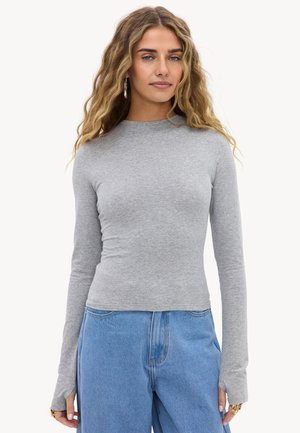 Vrouw met lang golvend haar, zilveren creolen, een strak lichtgrijs shirt met lange mouwen en duimgaten, en losse blauwe spijkerbroek.