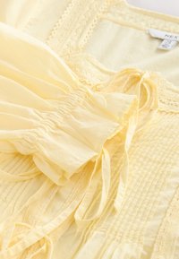 Jasnożółta bluzka o fakturowanej powierzchni, z marszczonymi rękawami, koronkowym wykończeniem i wiązaniami. Miękka tkanina o luźnym kroju.