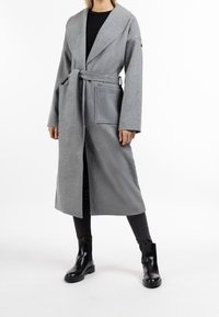 Manteau long gris en tissu texturé avec taille ceinturée, large col et deux poches avant. Porté par-dessus un pantalon noir et des bottes.