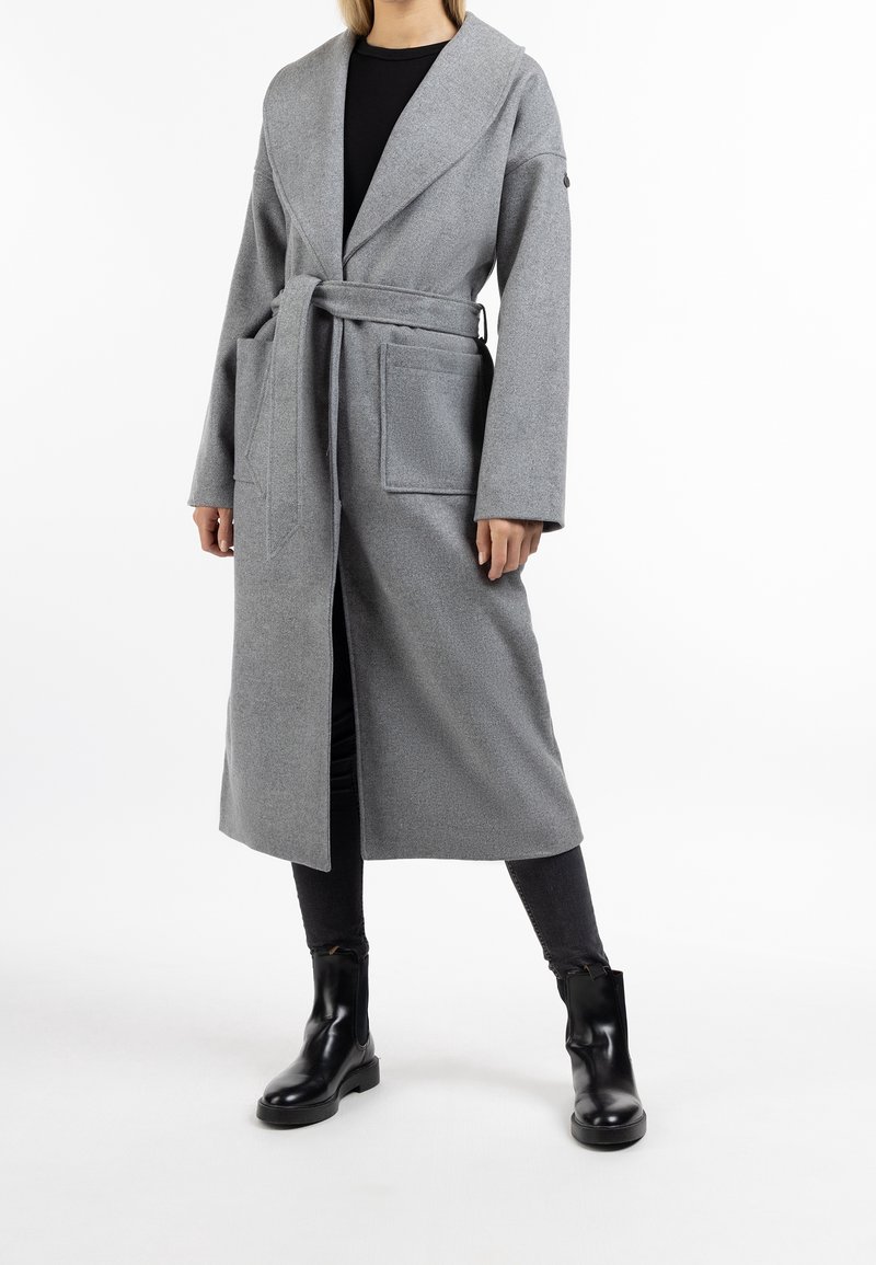 Manteau long gris en tissu texturé avec taille ceinturée, large col et deux poches avant. Porté par-dessus un pantalon noir et des bottes.