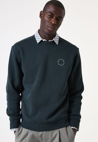 Sudadera verde oscura con cuello redondo, que presenta un detalle de logotipo circular. Debajo, se ve el cuello de una camisa blanca con un patrón. Se aprecian pantalones grises.