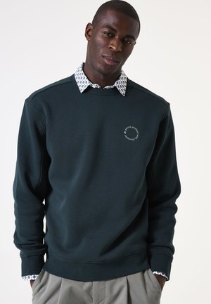 Sudadera verde oscura con cuello redondo, que presenta un detalle de logotipo circular. Debajo, se ve el cuello de una camisa blanca con un patrón. Se aprecian pantalones grises.