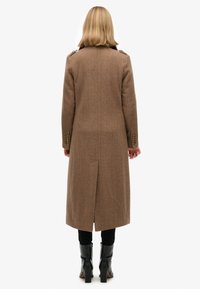 Brązowy płaszcz w jodełkę z akcentami na ramionach, długą sylwetką i rozcięciem po boku, noszony na czarnych spodniach i wysokich butach na obcasie.