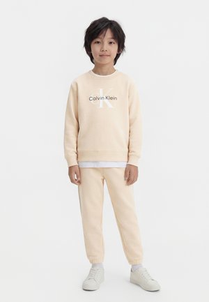 Barn står oppreist, iført beige Calvin Klein-genser, matchende joggebukser, hvite joggesko og en hvit undertrøye mot en ensfarget bakgrunn.