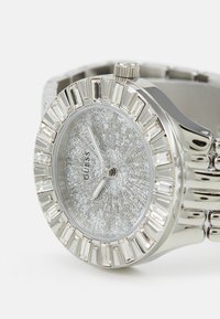 Zilveren roestvrijstalen horloge met een gestructureerde glinsterende wijzerplaat, kristalaccenten en een ronde bezel met rechthoekige details.