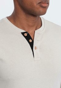 Camicetta henley a maniche corte color beige chiaro con patta nera e chiusura a collo con tre bottoni in legno. Tessuto morbido.