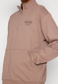 Světlý hnědý mikina Reebok se čtvrt zipem. Má měkkou tkaninu, žebrované manžety a přední kapsu. Logo vyšívané na hrudi.