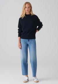 Pull en tricot bleu marine avec un ourlet et des poignets côtelés, associé à un jean droit bleu clair et des baskets blanches avec des accents bleus.