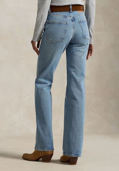 Személy világoskék egyenes szárú farmert, barna övet, barna magassarkú bokacsizmát és világosszürke hosszú ujjú felsőt visel, háttal a kamerának áll.