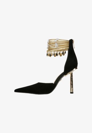 Zwarte suède pumps met spitse neus en een gouden en parel enkelband, voorzien van hangende zwarte en gouden accenten en een gestructureerde gouden hak.