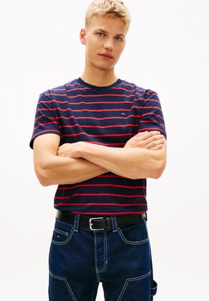 Jeune homme aux cheveux blonds courts portant un t-shirt rayé bleu marine et rouge et un jean bleu foncé avec une ceinture noire, les bras croisés.