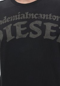 Μαύρο T-shirt με μεγάλο ξεθωριασμένο γκρι κείμενο που γράφει "DIESEL" και μικρότερο μερικώς ορατό κείμενο πάνω από το λογότυπο.
