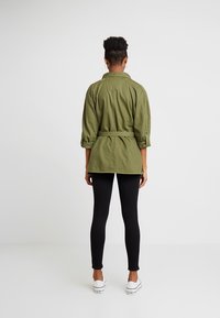Olive green button-up jacket with rolled sleeves and a belted waist, worn over black leggings and white sneakers. Smooth fabric texture.