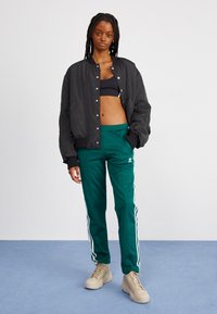 Giacca bomber nera sopra un crop top nero, abbinata a pantaloni della tuta verdi con strisce bianche e sneakers alte color sabbia.